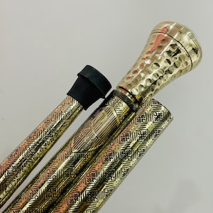 Op de afbeelding: Een inklapbare wandelstok met een gepolijst goudkleurig metalen handvat met een gehamerde textuur. De schacht is versierd met een ingewikkeld herhalend geometrisch patroon in goud- en kopertinten, voorzien van de gegraveerde tekst "R Artis".