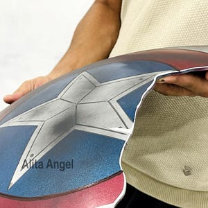K&ouml;nnte beinhalten: Ein Captain America Schild-Requisite mit einem Stern in der Mitte in den Farben Rot, Wei&szlig; und Blau. Das Schild hat eine metallische Oberfl&auml;che und einen Used-Look. Das Schild wird von einer Person gehalten.