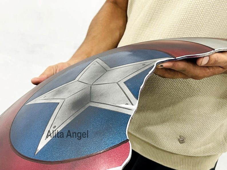 K&ouml;nnte beinhalten: Ein Captain America Schild-Requisite mit einem Stern in der Mitte in den Farben Rot, Wei&szlig; und Blau. Das Schild hat eine metallische Oberfl&auml;che und einen Used-Look. Das Schild wird von einer Person gehalten.