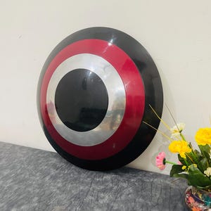 May include: A round shield with a black center, surrounded by silver, red, and black concentric circles. The shield is leaning against a white wall. A vase of flowers is in the background.