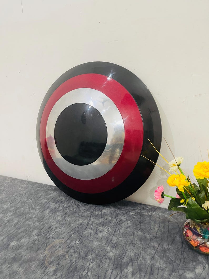 May include: A round shield with a black center, surrounded by silver, red, and black concentric circles. The shield is leaning against a white wall. A vase of flowers is in the background.