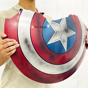 K&ouml;nnte beinhalten: Eine Captain America Schild-Requisite mit einem rot-wei&szlig;-blauen Farbschema. Das Schild hat einen Stern in der Mitte und ein abgenutztes, kampferprobtes Aussehen. Der Text "Alita Angel" ist auf dem Schild sichtbar.