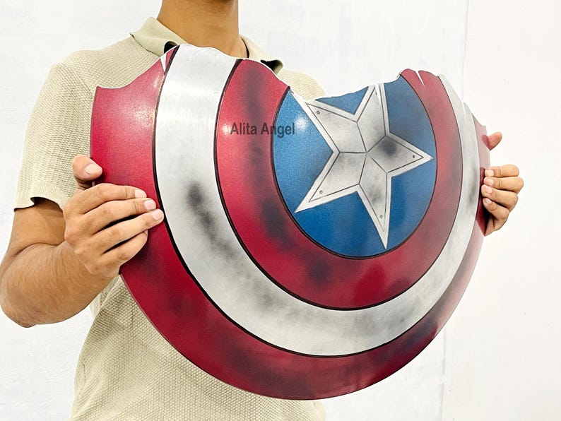 K&ouml;nnte beinhalten: Eine Captain America Schild-Requisite mit einem rot-wei&szlig;-blauen Farbschema. Das Schild hat einen Stern in der Mitte und ein abgenutztes, kampferprobtes Aussehen. Der Text "Alita Angel" ist auf dem Schild sichtbar.