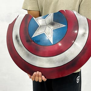 K&ouml;nnte beinhalten: Ein Captain America Schild-Requisite mit einem rot-wei&szlig;-blauen Farbschema und einem Sternemblem in der Mitte. Der Schild hat ein abgenutztes Aussehen. Der Text "Alita Angel" ist auf dem Schild sichtbar.