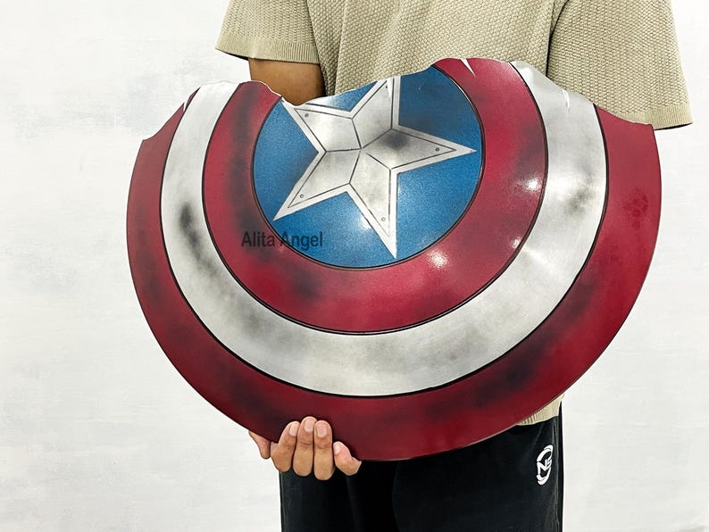 K&ouml;nnte beinhalten: Ein Captain America Schild-Requisite mit einem rot-wei&szlig;-blauen Farbschema und einem Sternemblem in der Mitte. Der Schild hat ein abgenutztes Aussehen. Der Text "Alita Angel" ist auf dem Schild sichtbar.