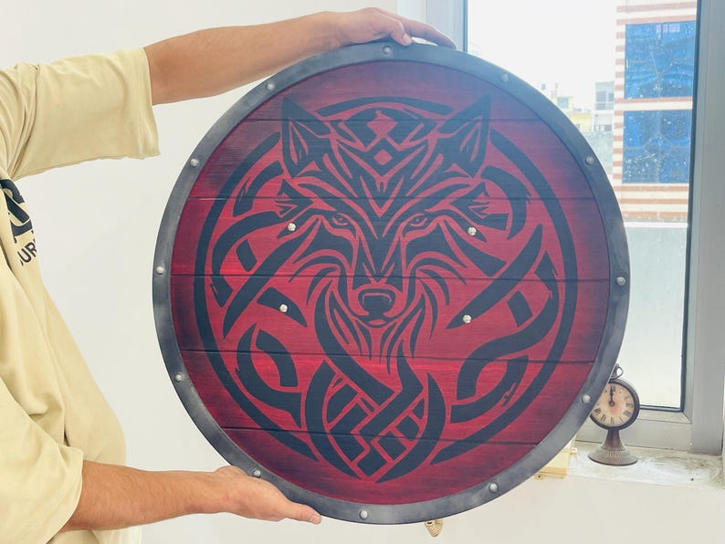 May include: Round wooden shield with a red background and a black wolf design. The shield has a metal rim and is held by a person. The wolf design is in the center of the shield.