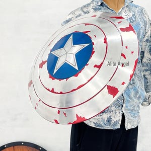 May include: A silver-colored shield with a blue central emblem featuring a white star. The shield has red accents and a circular design. The shield is a prop or collectible item.