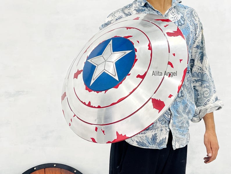 May include: A silver-colored shield with a blue central emblem featuring a white star. The shield has red accents and a circular design. The shield is a prop or collectible item.
