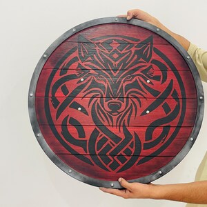 May include: Round wooden shield with a red background and a black wolf design. The shield has a metal rim and is held by a person. The wolf design is intricate and stylized, with a focus on the animal's face.