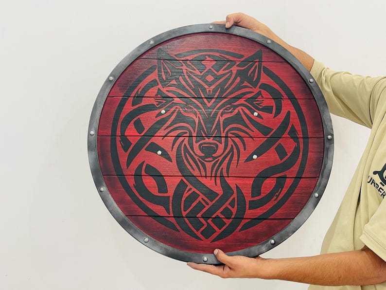 May include: Round wooden shield with a red background and a black wolf design. The shield has a metal rim and is held by a person. The wolf design is intricate and stylized, with a focus on the animal's face.