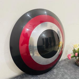 May include: A round shield with a black outer ring, a red middle ring, and a silver inner ring. The center of the shield is black. The shield is leaning against a wall.