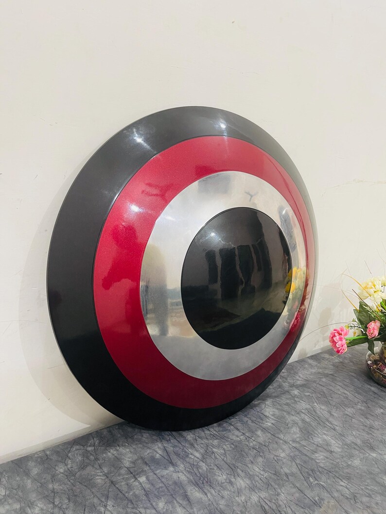 May include: A round shield with a black outer ring, a red middle ring, and a silver inner ring. The center of the shield is black. The shield is leaning against a wall.