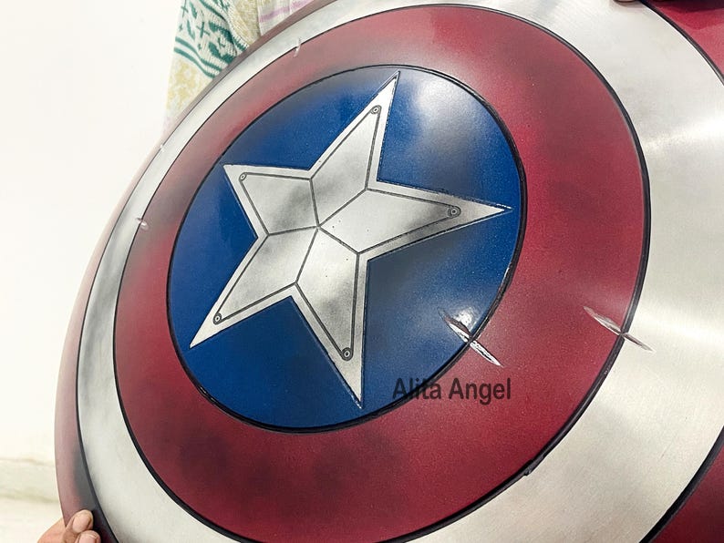 May include: A Captain America shield with a red, white, and blue color scheme. The shield features a white star on a blue center, surrounded by concentric red and silver rings. The shield has a distressed appearance with visible scratches.