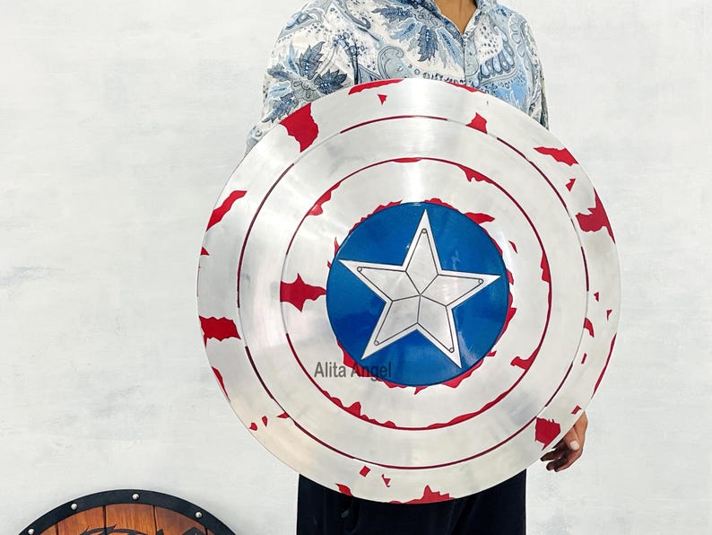 May include: A silver-colored shield with red accents and a blue circle featuring a white star in the center. The shield has a distressed appearance, with visible wear and tear. The shield is a replica of Captain America's shield.