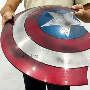 K&ouml;nnte beinhalten: Ein Captain America Schild-Requisite mit einem Used-Look-Design. Das Schild hat ein rot-wei&szlig;-blaues Farbschema mit einem zentralen Stern-Emblem. Das Schild wird von einer Person gehalten.
