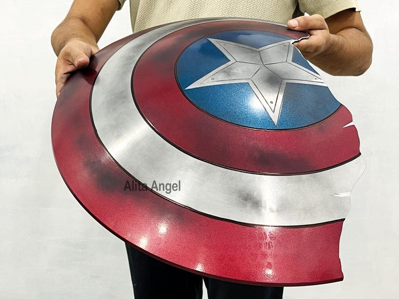 K&ouml;nnte beinhalten: Ein Captain America Schild-Requisite mit einem Used-Look-Design. Das Schild hat ein rot-wei&szlig;-blaues Farbschema mit einem zentralen Stern-Emblem. Das Schild wird von einer Person gehalten.