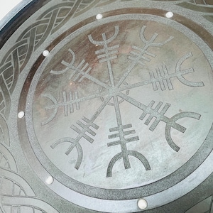 May include: Close-up of a circular, metallic surface with a detailed Viking-inspired design. The central focus is the Vegv&iacute;sir symbol, surrounded by intricate knotwork and small, evenly spaced circular accents. The color is a muted gray.
