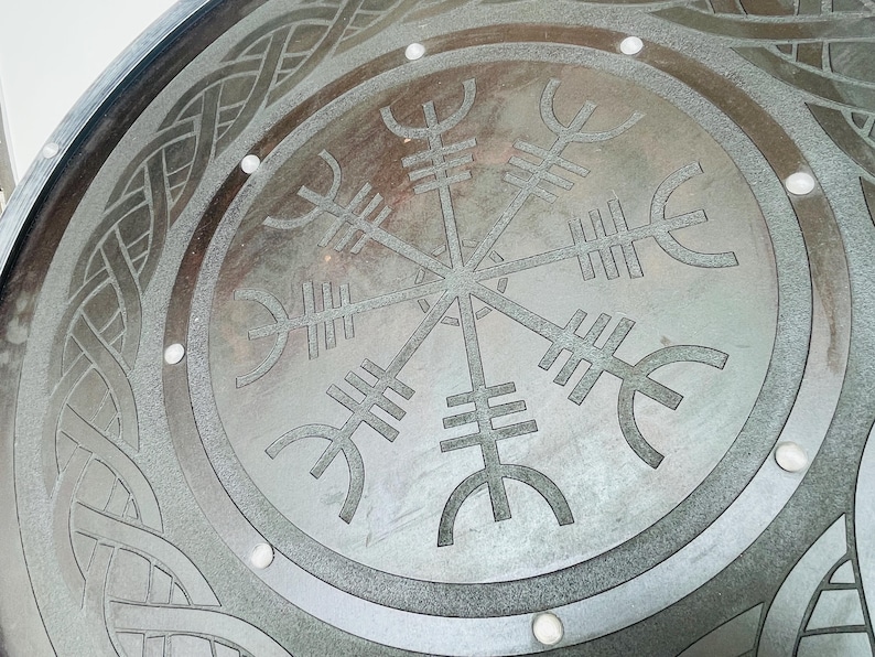 May include: Close-up of a circular, metallic surface with a detailed Viking-inspired design. The central focus is the Vegv&iacute;sir symbol, surrounded by intricate knotwork and small, evenly spaced circular accents. The color is a muted gray.
