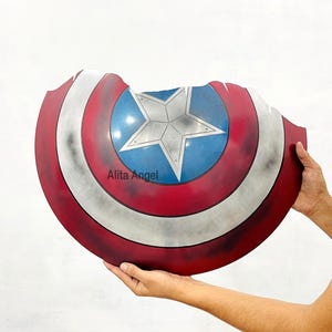 K&ouml;nnte beinhalten: Eine Captain America Schild-Requisite mit einem rot-wei&szlig;-blauen Farbschema und einem Sternemblem in der Mitte. Der Schild hat ein abgenutztes, kampferprobtes Aussehen. Der Text "Alita Angel" ist auf dem Schild zu sehen.