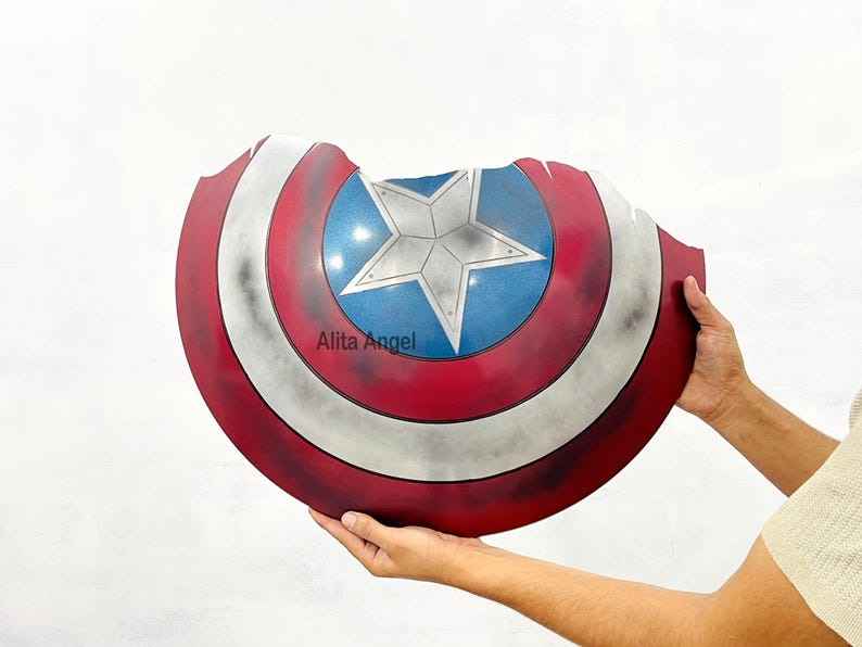 K&ouml;nnte beinhalten: Eine Captain America Schild-Requisite mit einem rot-wei&szlig;-blauen Farbschema und einem Sternemblem in der Mitte. Der Schild hat ein abgenutztes, kampferprobtes Aussehen. Der Text "Alita Angel" ist auf dem Schild zu sehen.