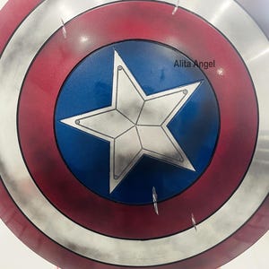 May include: A round shield with a red, white, and blue color scheme. The shield features a central blue circle with a white star, surrounded by concentric rings of red, silver, and red. The text "Alita Angel" is visible on the shield.