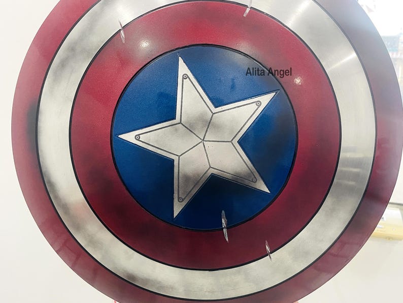 May include: A round shield with a red, white, and blue color scheme. The shield features a central blue circle with a white star, surrounded by concentric rings of red, silver, and red. The text "Alita Angel" is visible on the shield.