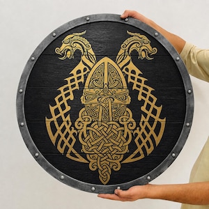 May include: A circular wooden shield featuring a black finish with a gold-colored intricate knotwork design of a central helmet flanked by two dragon heads. The shield has a metallic gray rim secured with evenly spaced silver-toned rivets.
