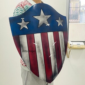以下が含まれることがあります： 3つの白い星と赤と白のストライプが特徴の青い上部セクションを備えたシールド。シールドは湾曲した形状で、下部には「Alita Angel」の文字が見えます。
