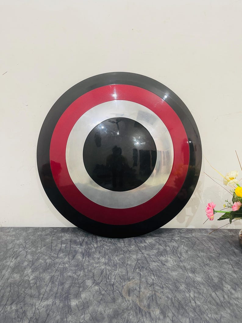 May include: A round shield with a black, red, white, and silver color scheme. The shield has a central black circle, surrounded by a silver ring, a red ring, and a black outer rim. The shield is displayed against a neutral background.