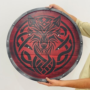 Op de afbeelding: Ronde houten schild met een rode achtergrond en een zwart wolfontwerp. Het schild heeft een metalen rand en wordt door handen vastgehouden. Het wolfontwerp is een gestileerd tribaal patroon.