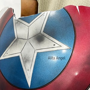 K&ouml;nnte beinhalten: Eine Nachbildung des Captain America-Schildes mit einem zentralen blauen Kreis mit einem wei&szlig;en Stern, umgeben von roten und wei&szlig;en Ringen. Auf dem Schild steht der Text "Alita Angel" auf dem blauen Kreis.