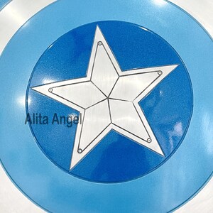 May include: A circular shield featuring a white star emblem at its core, encircled by bands of blue and white. The shield has a metallic finish, with the text "Alita Angel" present.