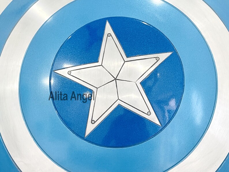 May include: A circular shield featuring a white star emblem at its core, encircled by bands of blue and white. The shield has a metallic finish, with the text "Alita Angel" present.