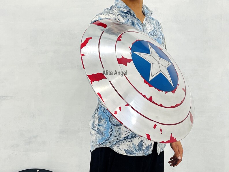 May include: A silver and red Captain America shield with a blue star emblem. The shield has a distressed finish and is round in shape. The shield is a prop or collectible item.