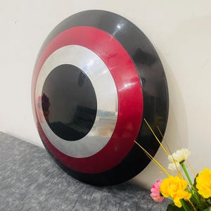 May include: A round shield with a black center, a silver ring, a red ring, and a black outer edge. The shield is leaning against a white wall. A bouquet of yellow and white flowers is in the foreground.