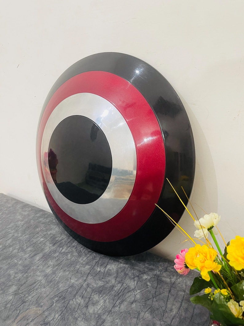 May include: A round shield with a black center, a silver ring, a red ring, and a black outer edge. The shield is leaning against a white wall. A bouquet of yellow and white flowers is in the foreground.