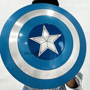 May include: A blue, silver, and white shield with a central white star design. The shield has a circular shape with concentric rings of colour. The shield is a prop or costume accessory.
