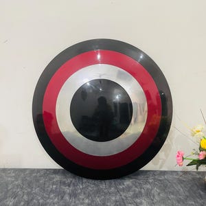 May include: A round shield with a black outer ring, a red band, a silver band, and a black center. The shield is a replica of a superhero's shield.