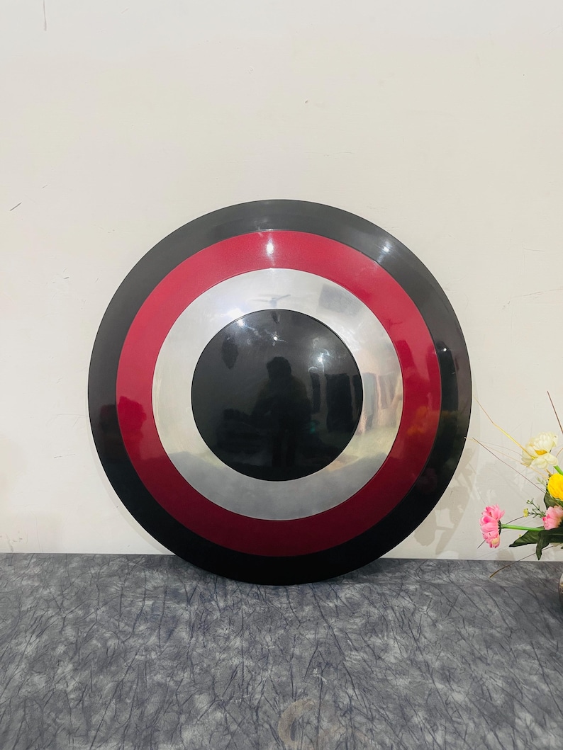 May include: A round shield with a black outer ring, a red band, a silver band, and a black center. The shield is a replica of a superhero's shield.