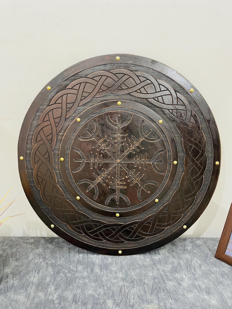 May include: A round, dark brown wooden shield with intricate carved knotwork and a central compass-like symbol. The shield is adorned with small, gold-colored studs around the perimeter, showcasing a Viking or Norse theme.