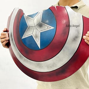 K&ouml;nnte beinhalten: Eine Captain America Schild-Requisite mit einem Used-Design in Rot, Wei&szlig; und Blau. Das Schild hat einen zentralen blauen Kreis mit einem wei&szlig;en Stern, umgeben von roten und wei&szlig;en Streifen. Das Schild wird von einer Person gehalten.
