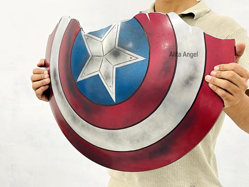 K&ouml;nnte beinhalten: Eine Captain America Schild-Requisite mit einem Used-Design in Rot, Wei&szlig; und Blau. Das Schild hat einen zentralen blauen Kreis mit einem wei&szlig;en Stern, umgeben von roten und wei&szlig;en Streifen. Das Schild wird von einer Person gehalten.