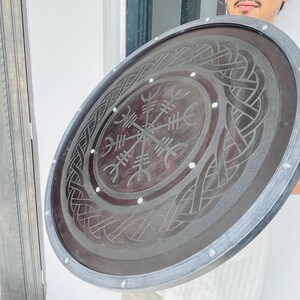 May include: A large, round shield with a dark brown center and a silver-colored rim. The shield features an intricate, engraved design with a central compass-like symbol and interwoven patterns. Small, white rivets secure the rim.