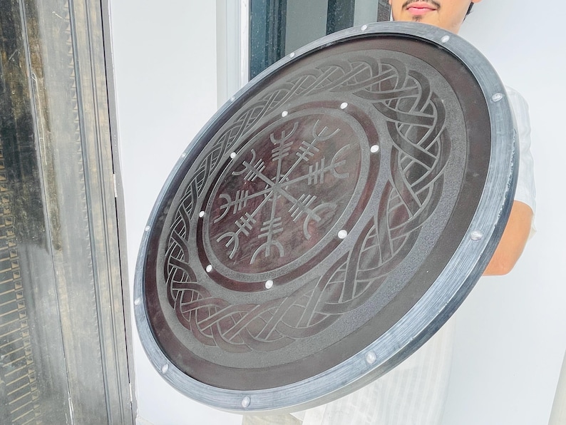 May include: A large, round shield with a dark brown center and a silver-colored rim. The shield features an intricate, engraved design with a central compass-like symbol and interwoven patterns. Small, white rivets secure the rim.