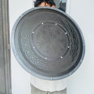 May include: A large, round, dark gray shield with intricate Celtic knot and rune designs. The shield has a silver-colored trim and rivets around the edge. The central design features a compass-like symbol. A decorative item.