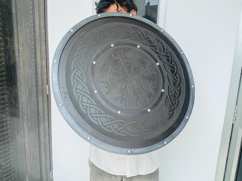 May include: A large, round, dark gray shield with intricate Celtic knot and rune designs. The shield has a silver-colored trim and rivets around the edge. The central design features a compass-like symbol. A decorative item.