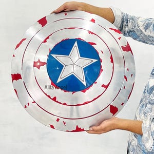 May include: A silver-colored shield with a blue circle in the center, featuring a white star. The shield has red accents around the edges and is held by a person. The shield is a replica of Captain America's shield.