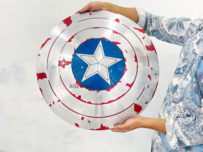 May include: A silver-colored shield with a blue circle in the center, featuring a white star. The shield has red accents around the edges and is held by a person. The shield is a replica of Captain America's shield.