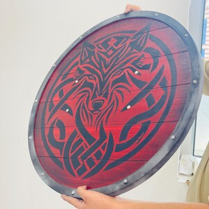 May include: A round wooden shield with a red background and a black wolf design. The shield has a metal rim and rivets. The wolf design is a stylized tribal illustration.
