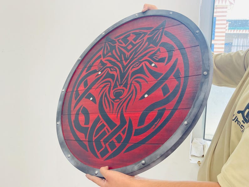May include: A round wooden shield with a red background and a black wolf design. The shield has a metal rim and rivets. The wolf design is a stylized tribal illustration.